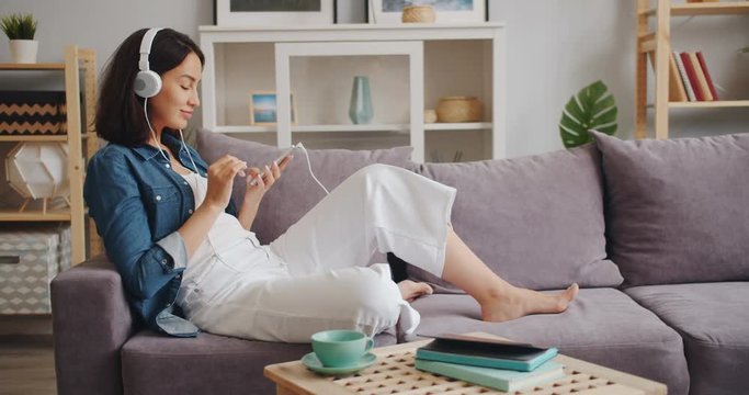 Joyful young lady in trendy clothing is enjoying music in headphones and touching smartphone screen at home. Modern technology and happy youth concept.