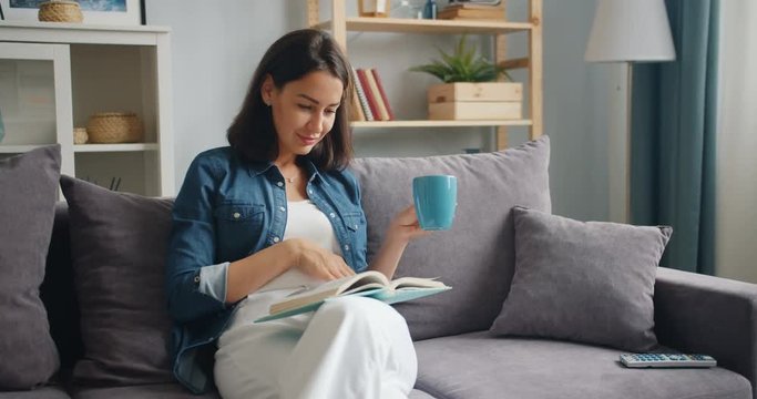 Smart young lady in casual clothing is reading book holding cup of tea sitting on couch in apartment relaxing. Millennials, modern lifestyle and house concept.