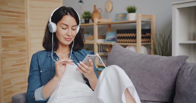 Beautiful young woman is listening to music through headphones using smartphone smiling relaxing in apartment. Millennials, house and youth culture concept.