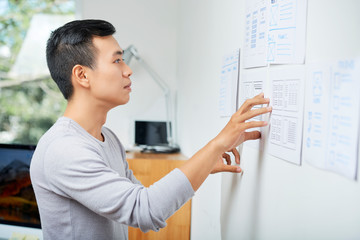 Pensive young Asian UX designer looking at documents with wireframes hanging on wall in office