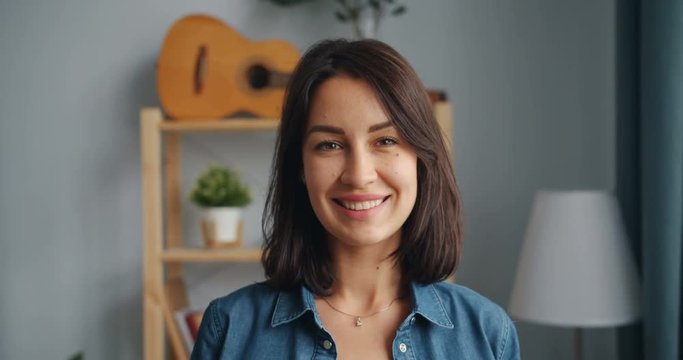 Slow motion portrait of charming young woman looking at camera smiling standing at home alone. Cosy apartment with furniture and guitar visible in background.