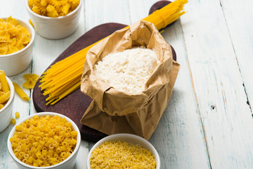 uncooked pasta variations and flour in paper bag, on white wooden table