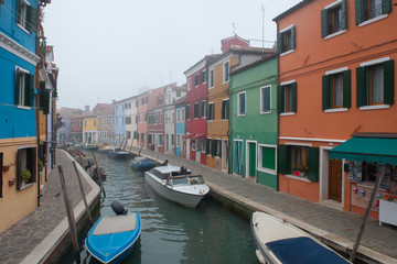 Fototapeta na wymiar Misty picture of street with colored houses in fog, italian island Burano, province of Venice, Italy, foggy weather. Little beautiful dock with boats, Mediterranean sea.