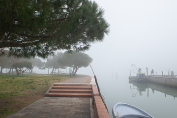 Wharf in quit bay, foggy weather, italian island Burano, province of Venice, Italy. Little beautiful dock with tree and boats in fog, Mediterranean sea.