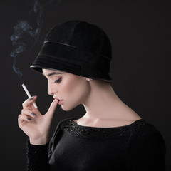 Obraz premium Young fashion woman smoking cigarette in hat and black drees over dark background. Vintage female portrait, styling. Image toned.