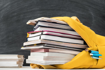 Information overload. Yellow backpack stuffed with books and note pad over empty chalkboard.