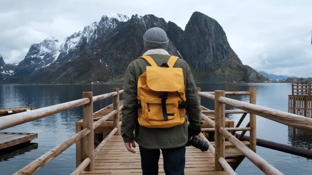 Traveler goes on a wooden pier. dressed in a jacket and knitted hat and a yellow backpack. back view. Norway lofoten
