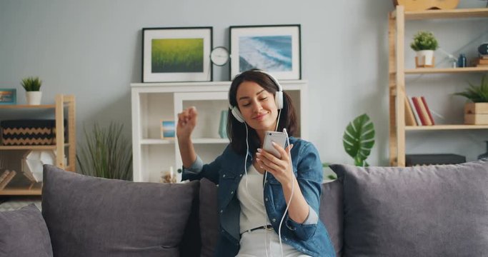 Happy relaxed girl is listening to music in headphones holding smartphone touching screen sitting on sofa at home. People, apartment and gadgets concept.