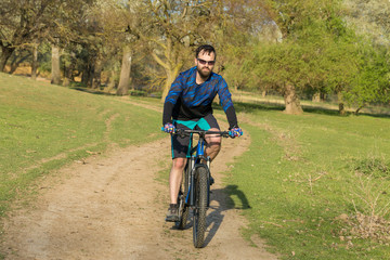 Obraz premium Rowerzysta w szortach i koszulce na nowoczesnym rowerze typu hardtail z włókna węglowego z widelcem pneumatycznym jeździ w terenie po zielonych wzgórzach w pobliżu lasu