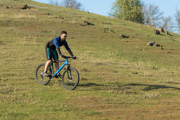 Cyclist in shorts and jersey on a modern carbon hardtail bike with an air suspension fork rides off-road on green hills near the forest	