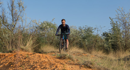 Cyclist in shorts and jersey on a modern carbon hardtail bike with an air suspension fork rides off-road on the orange-red hills at sunset evening in summer	
