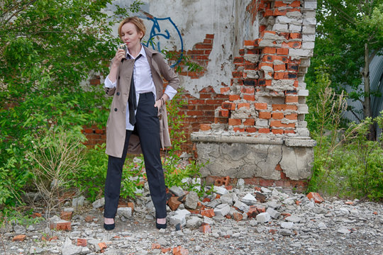Woman of androgynous appearance and short blonde hair bob hairstyle in a coat, men's white shirt, tie and trousers, smoking a cigarette with an abandoned ruins on background. Noir detective or police