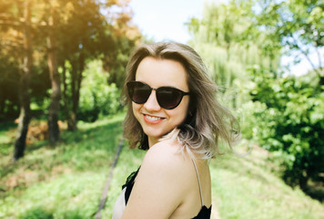 Young hipster girl in fashionable top is standing on railway tracks in the park. Summer photoshoot outdoors for lifestyle concept. Blonde woman in cool glasses is waving hair.