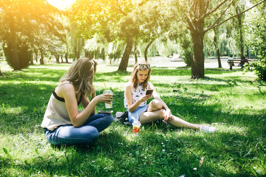 Two hipster girls are sitting on the grass with summer cocktails. Sisters are looking at their smartphones. Social networks communication. Cold non-alcoholic drinks with ice to go. Mojito and lemonade