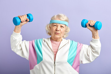 Studio shot of fitness mature female pensioner working out with dumbbells isolated over purple background