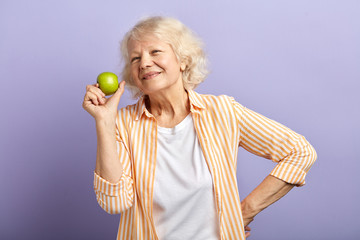 Mature age and healthy eating. Beautiful senior woman smiling satisfied, holding a a green apple, showing good food ifor old age, isolated on purple with copyspace.