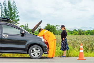 Repair technician and unfortunate female driver with broken down car. He is repairing it at roadside in rural area.