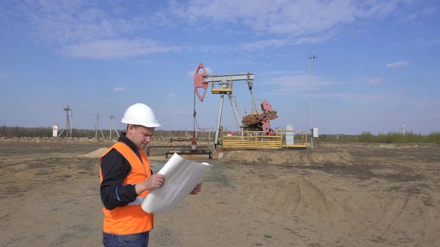 A young caucasian engineer in a signal vest and protective helmet looks at the diagram of the oil, gas and fuel pumping machine, the oil refining industry, supervisor