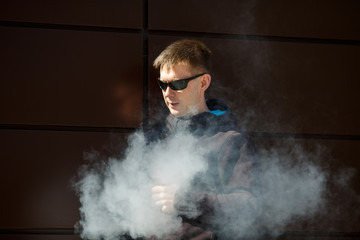 Vape man. An adult bearded man in sunglasses smokes an electronic cigarette on a sunny day outside. Bad habit that is harmful to health. Vaping aclivity.