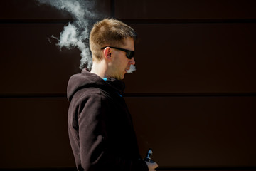 Fototapeta na wymiar Vape man. An adult bearded man in sunglasses smokes an electronic cigarette on a sunny day outside. Bad habit that is harmful to health. Vaping aclivity.