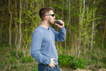 Vape man. An adult white bearded man in glasses smokes an electronic cigarette outside in the forest in sunny day. Bad habit that is harmful to health.