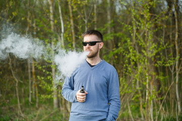 Vape man. An adult white bearded man in glasses smokes an electronic cigarette outside in the forest in sunny day. Bad habit that is harmful to health.