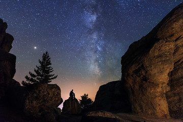 Naklejka na ściany i meble Man sitting under the stars Milky Way in the mountains silhouette rock.