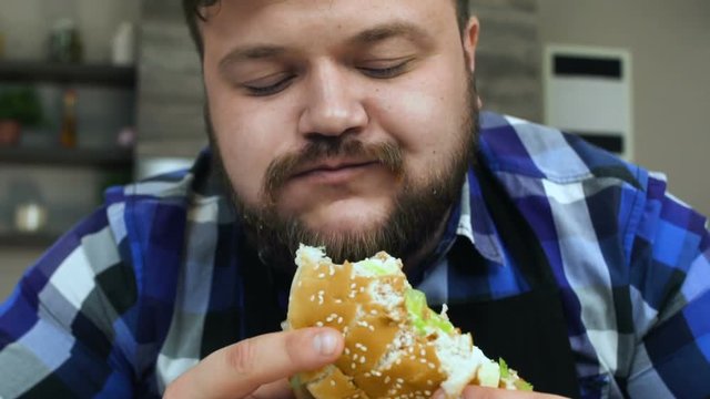 Fat man with a beard bites a piece of hamburger. The food is very delicious. The plump guy is glad. Unhealthy Lifestyle, fried and high calorie food. The risk of obesity and overweight. Slow motion