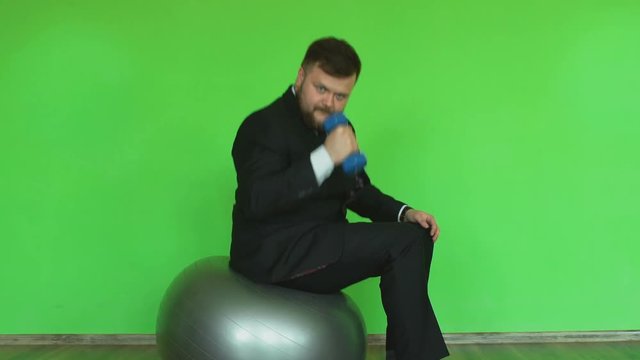 Funny plump man in suit with dumbbell sits and moves on Exercise ball in fitness club. Chromakey Green. Bearded thick guy in black jacket, shirt and tie is in gym