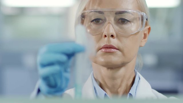 Rack focused shot of female doctor in special spectacles and gloves holding test tube with blue liquid and pipette in her hands and carrying out test with reagents