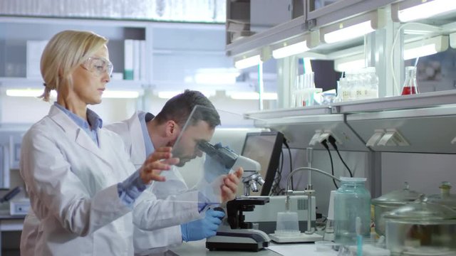 Medium shot of male scientist doing research with microscope and communicating to his female colleague standing nearby and using gadget with invisible AR screen
