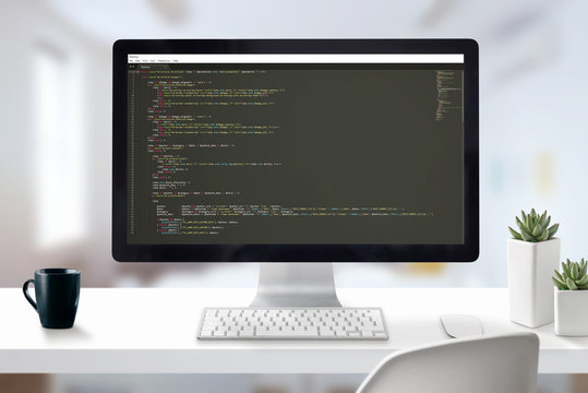 Web developer computer with javascript code on display. Work desk close-up scene with office in background.