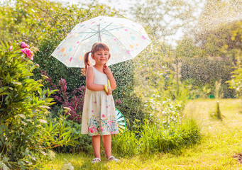 Happy little girl in garden under the summer rain with an umbrella.