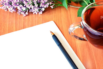 Blooming flower of purple lilac branch, glass tea Cup, black ballpoint pen and white blank sheet of paper with empty space for writing of text on wooden table