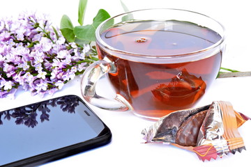 bouquet blooming flower of purple lilac branch, glass black tea Cup, sweet chocolate candy in shiny wrapper and black classic modern smartphone with reflection in display on white background