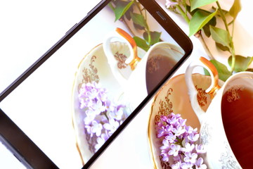 Elegant vintage Cup with golden handle of tea on saucer with beautiful violet lilac flowers on white background and Black classic modern smartphone with this picture on the screen. Tilt horizon frame