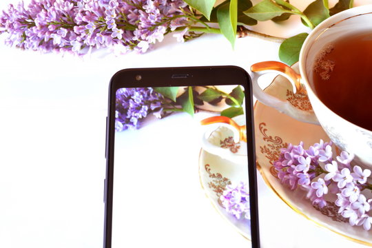 Elegant vintage Cup with golden handle of tea on saucer with beautiful violet lilac flowers on white background and Black classic modern smartphone with this picture on the screen. Horizontal frame