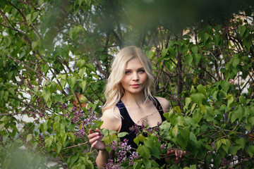 Sexy naughty blue-eyed light-skinned young blonde posing next to a Bush of blooming lilac. Soft focus, copy space