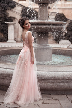 Beautiful bride in pink wedding dress. Outdoor romantic portrait of attractive brunette woman with hairstyle in prom dress with tulle skirt posing by fountain at park.