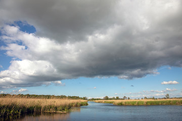 A ditch across the reed fields under a light blue sky with heavy rainy clouds in de Nieuwkoopse plassen in the Netherlands.