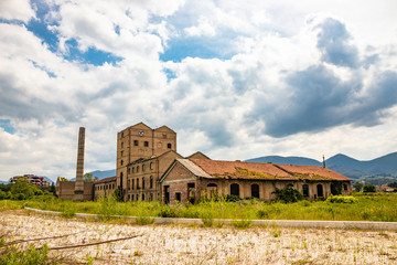 Fototapeta na wymiar An old disused factory, abandoned and in ruins, with a smashed roof and a chimney. Tall weeds invade the building. Concept of economic bankruptcy. Cloudy sky. Italy, Foligno, Umbria.