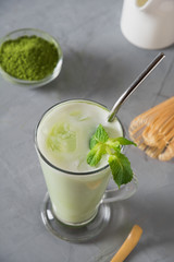 Green cold matcha tea in latte glass with ice cube and milk on grey table.