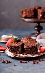 pieces of chocolate nut brownie, on saucer with slices of strawberries, crumbs. Blue, grey background.