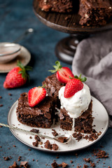 pieces of chocolate nut brownie, with scoop of ice cream, on white plate, spoon, with slices of strawberries, crumbs, grey textile. Dark blue background. Vertical