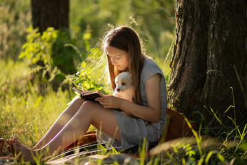 A girl sitting near a tree and reading a book, holding a labrador puppy. At sunset in the forest in...