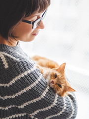 Cute ginger cat dozing on woman arms. Smiling woman in grey knitted sweater stroking her fluffy pet. Cozy home.