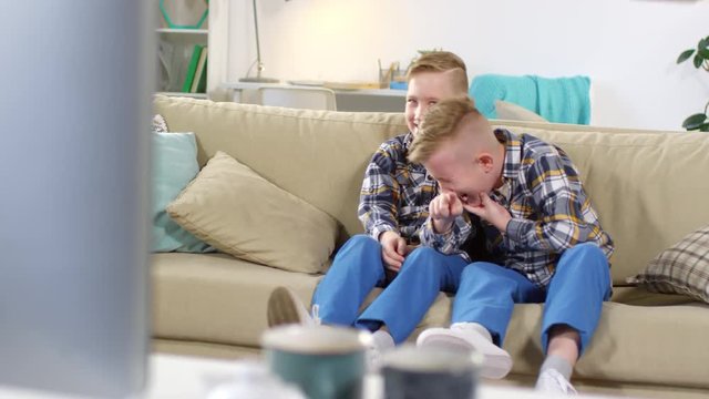 Full shot of 9-year-old blond Caucasian identical twins sitting on couch at home and watching something funny on TV, pointing towards screen and rolling with laughter
