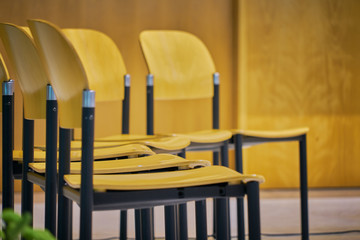 Rows of empty chairs prepared for an indoor event. Chairs made of light brown wood with black metal structure.