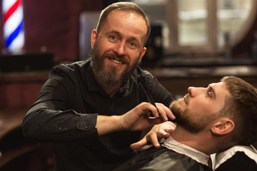 Front view of funny barber in black shirt looking at camera and smiling while doing trendy beard cut in barbershop. Skilled male master shaving beard with razor to client. Concept of grooming.