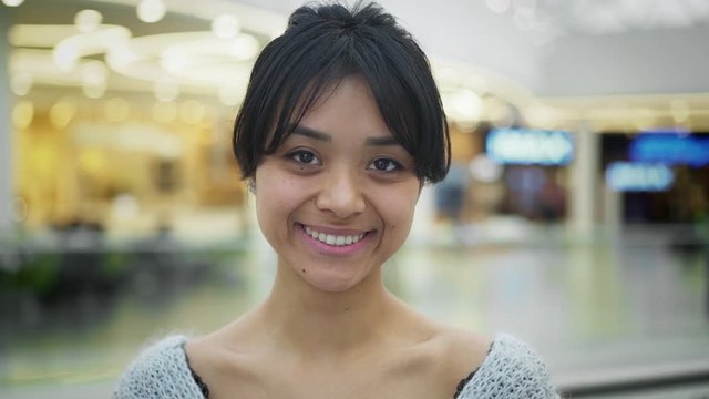 Portrait slow motion close up gimbal shot of smiling happy black hair asian female with beautiful smile wearing grey sweatshirt standing on background of mall